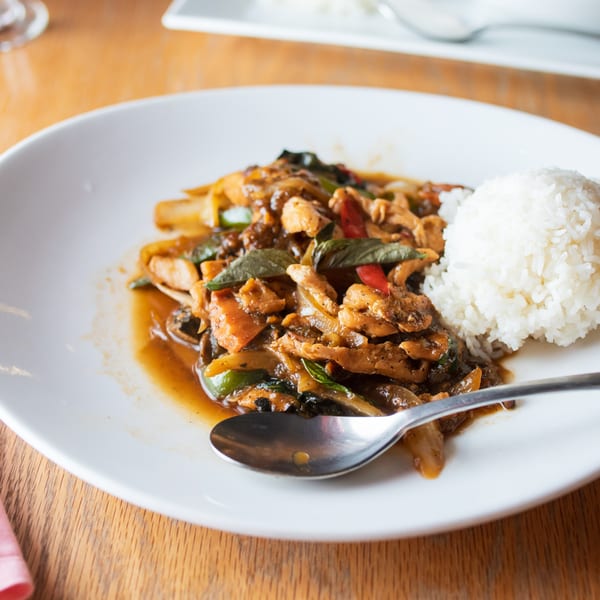 A white plate of Thai basil chicken with rice and a spoon on a wooden table.