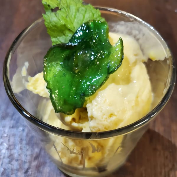 Scoops of yellow ice cream in a glass, garnished with fresh mint and a candied green leaf.