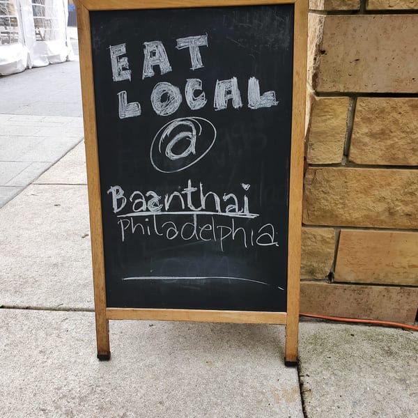 Chalkboard sign on a sidewalk reads "EAT LOCAL @ Baanthai Philadelphia" in white chalk.
