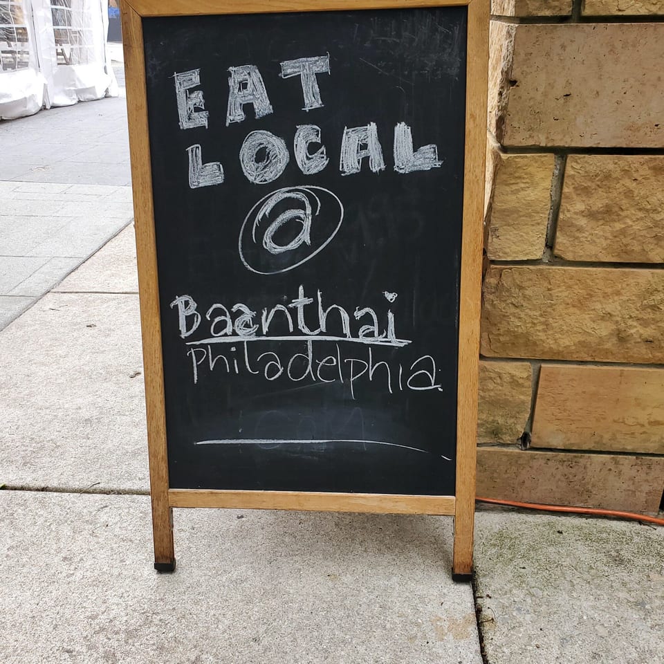 Chalkboard sign on a sidewalk reads "EAT LOCAL @ Baanthai Philadelphia" in white chalk.