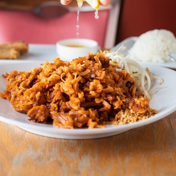A hand squeezing lime juice over a plate of Baan Thai's Pad Thai noodles with bean sprouts and rice.