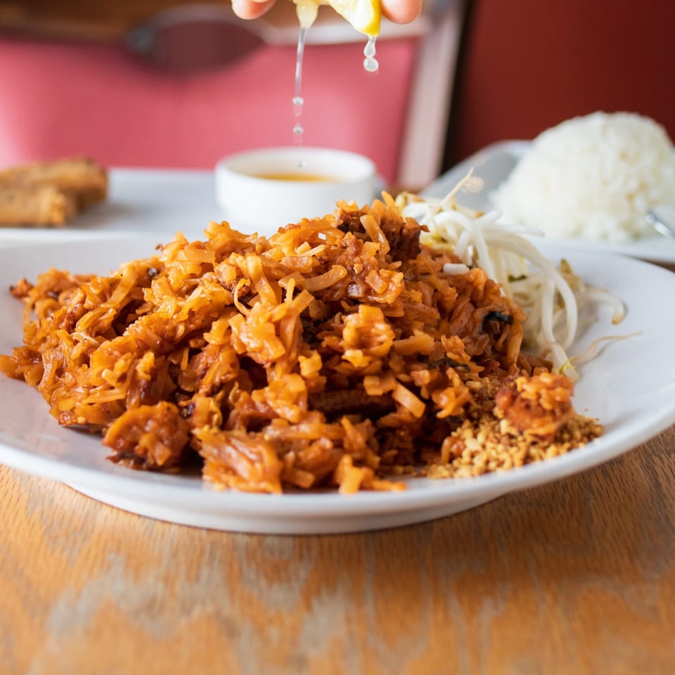 A hand squeezing lime juice over a plate of Baan Thai's Pad Thai noodles with bean sprouts and rice.